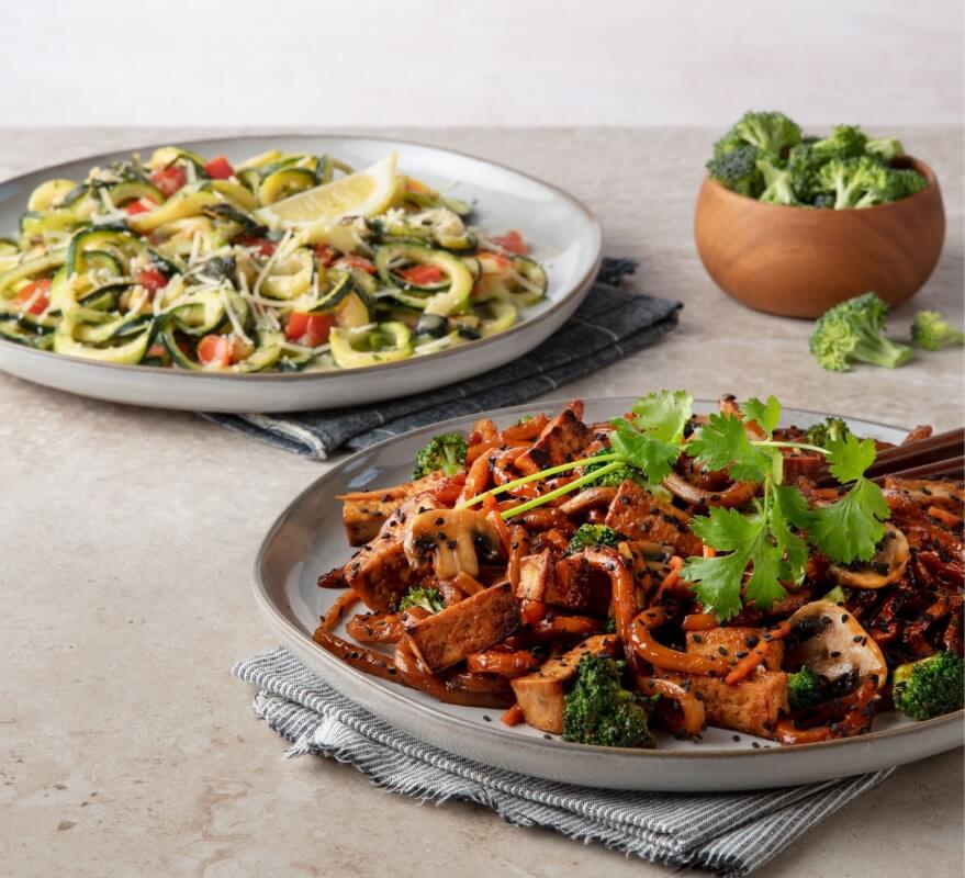 two plates with noodles dishes and a bowl of broccoli beside them