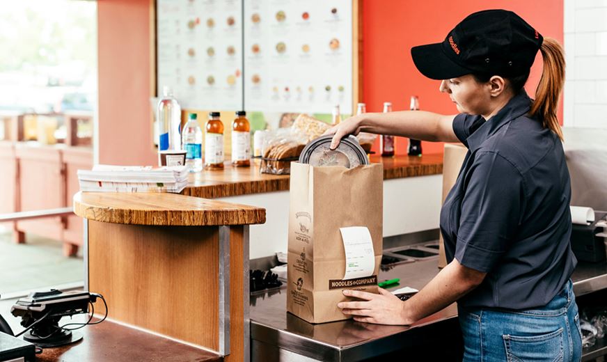 Noodles & Company franchise employee places noodle dish in to-go bag at front counter of restaurant