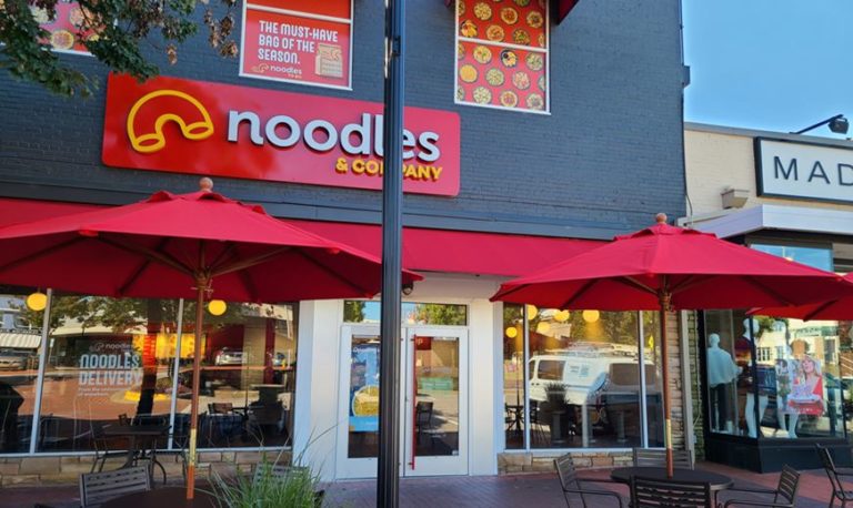 Exterior of a Noodles & Company franchise with red umbrellas and patio seating
