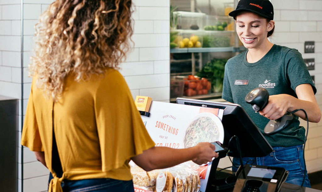 Noodles & Company franchise team member scans loyalty app for customer in yellow blouse