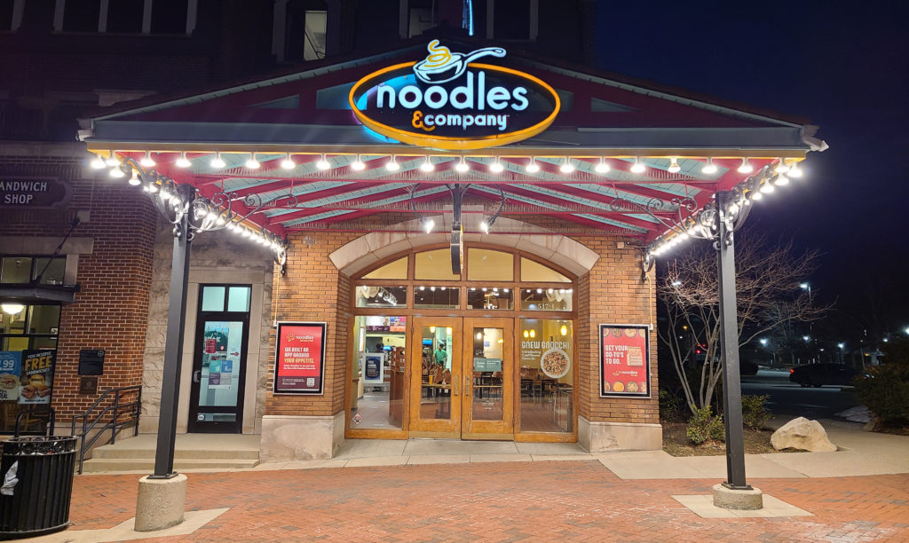 A picture of the exterior of a Noodles & Company franchise at night, with a string of lights on the awning