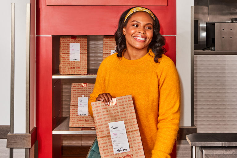 Woman standing and smiling with a Noodles to-go brown bag.